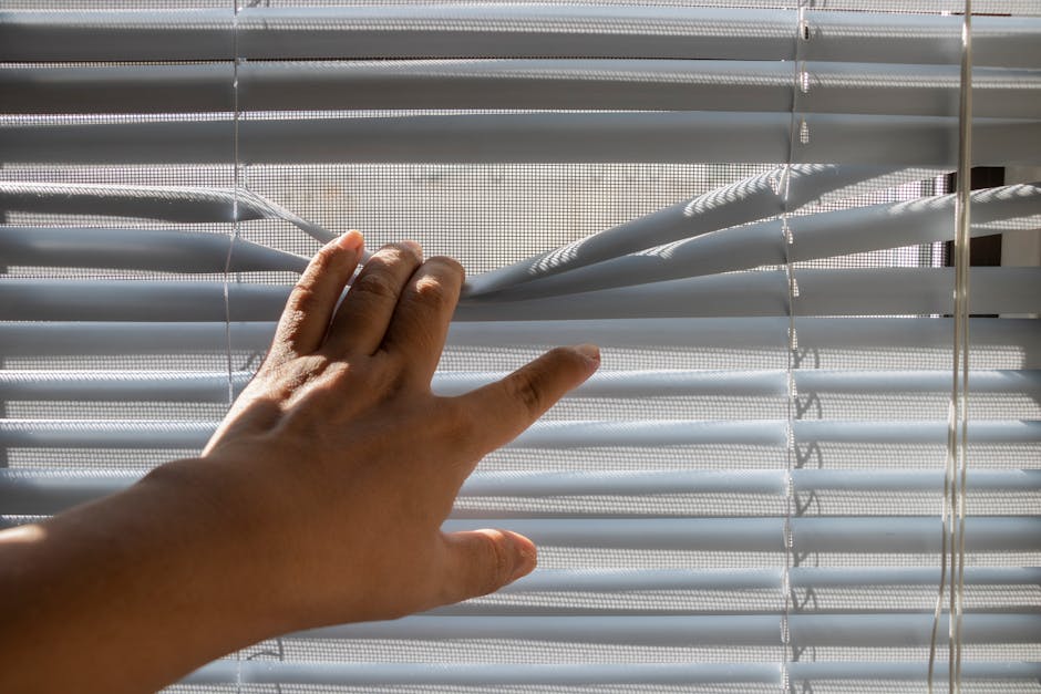 A hand gently opens window blinds, letting sunlight stream indoors.