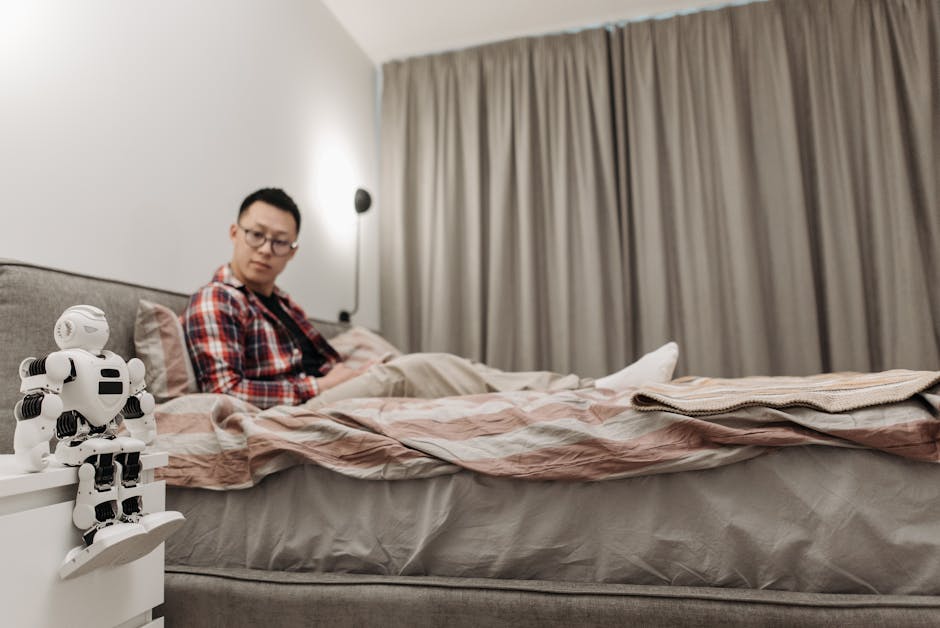 Asian man sitting in bedroom, observing futuristic robot on nightstand.