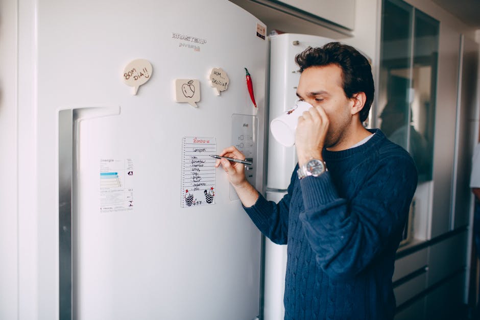 Young male employee in casual outfit drinking coffee and taking notes on paper attached on fridge in morning
