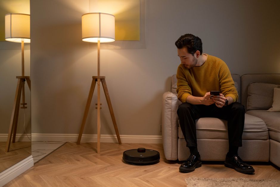 Adult man with a smartphone monitors robot vacuum in a modern living room.