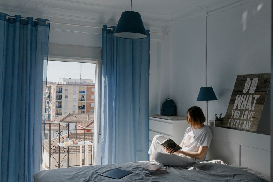 Woman in a white shirt enjoys a book, sitting in a sunlit bedroom near a window with blue curtains.