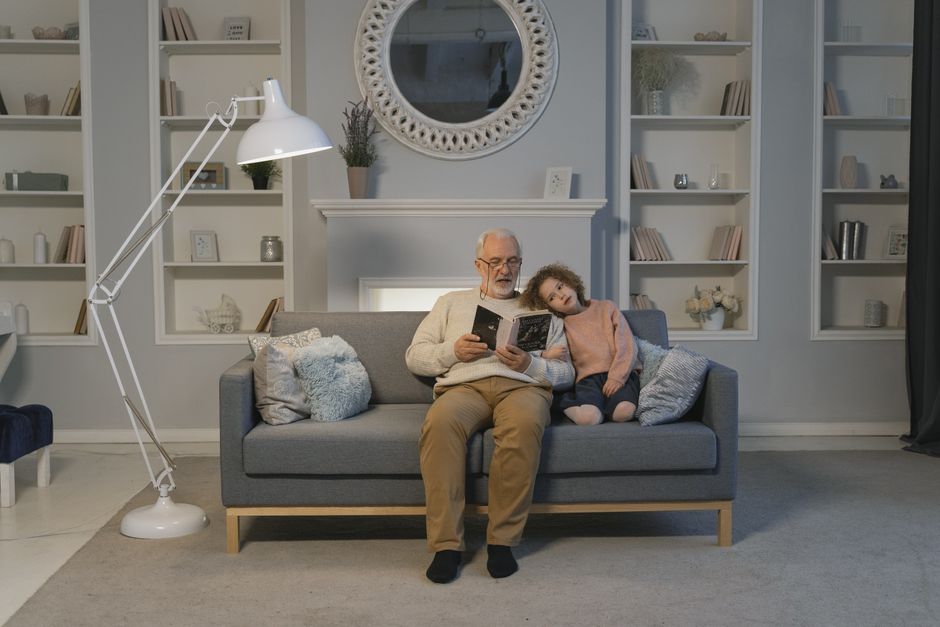 A cozy scene with a grandfather reading to his granddaughter at home on the sofa.
