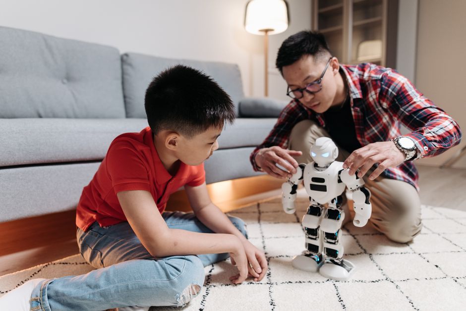 A father and son engage with a humanoid robot on a living room carpet, fostering creativity.