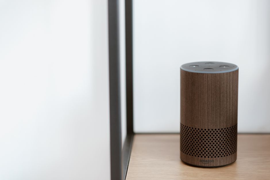 A sleek, modern smart speaker displayed on a wooden shelf in a minimalist room setting.