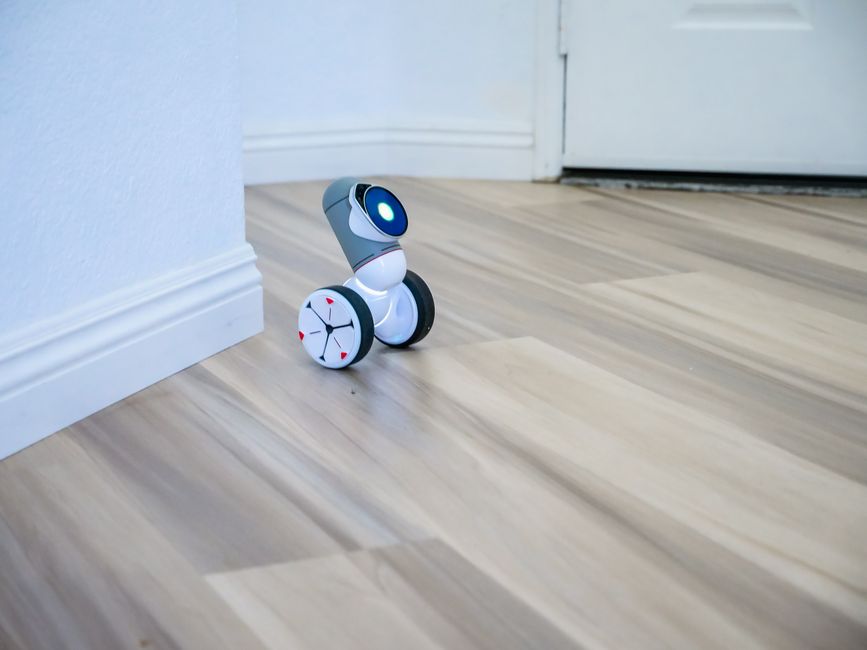 A small wheeled robot with a digital face navigating a wooden floor indoors.