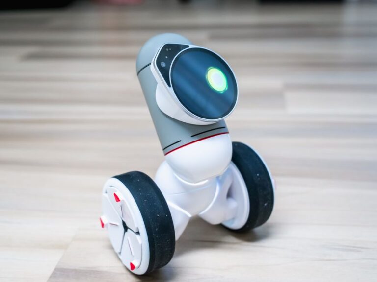 A small wheeled robot with a glowing light on a wooden floor indoors.