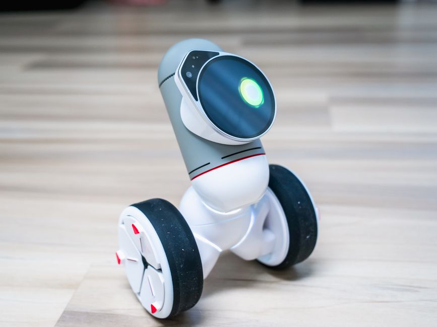 A small wheeled robot with a glowing light on a wooden floor indoors.