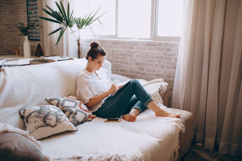 Adult woman sitting on sofa doing remote work on tablet in cozy living room.