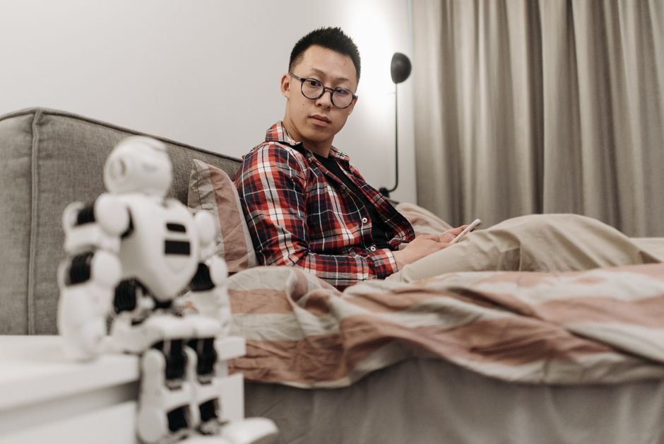 Asian man in plaid shirt sitting on bed, observing a robot toy in a cozy bedroom setting.