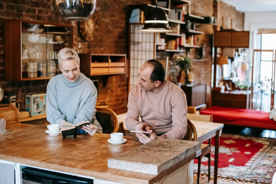 Concentrated adult ethnic man browsing tablet and drinking coffee while sitting at table near smiling wife reading interesting book during weekend at home