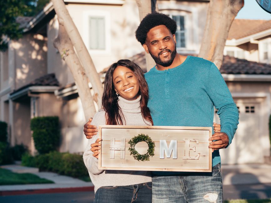 Happy couple holding a 'Home' sign in front of their new house, symbolizing joy and togetherness.