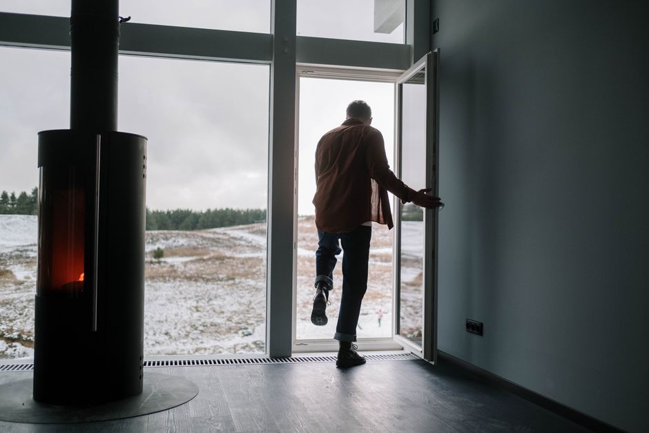 Man exiting modern home onto snowy landscape through glass door with a fireplace inside.