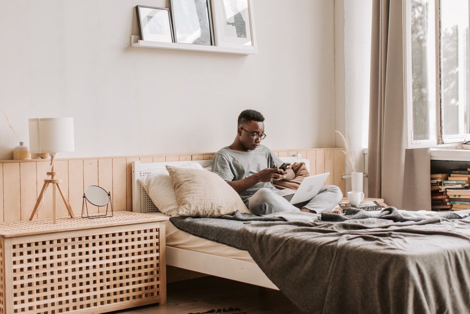 Man in comfortable bedroom using smartphone and laptop for remote work, surrounded by soft decor.