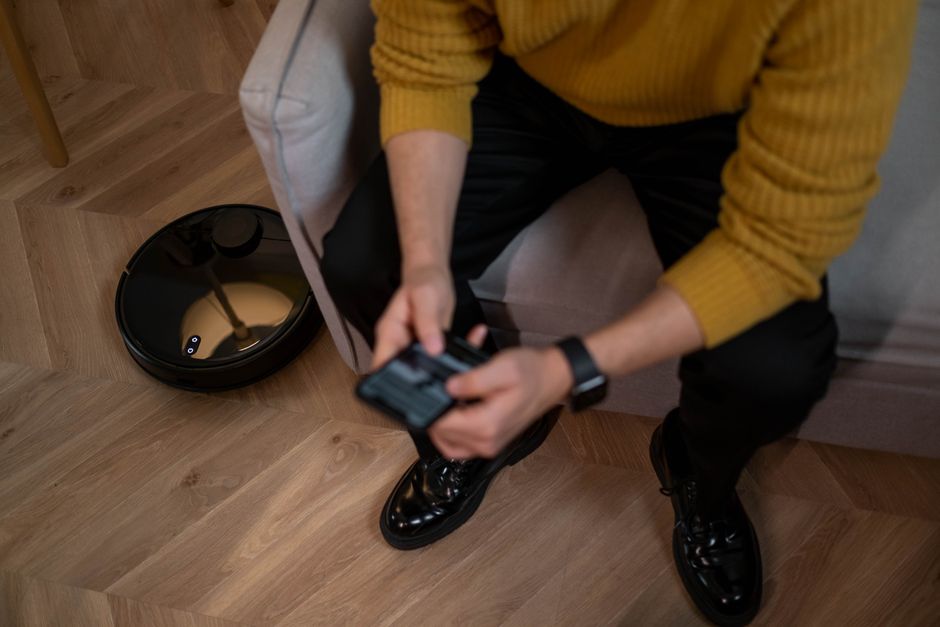 Man in yellow sweater using remote to control robotic vacuum on wooden floor.