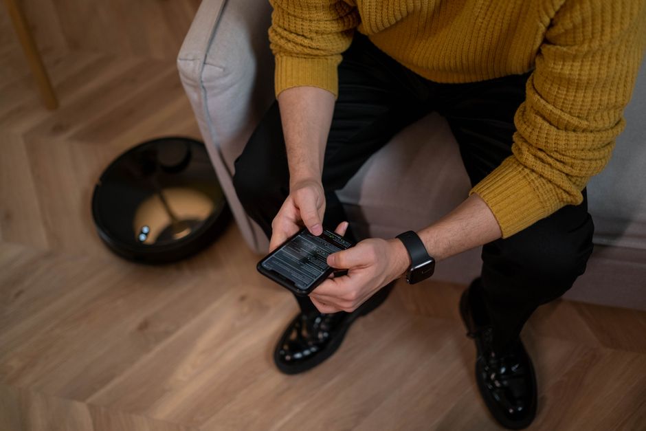 Man in yellow sweater using smartphone, robotic vacuum on wooden floor, modern home setting.