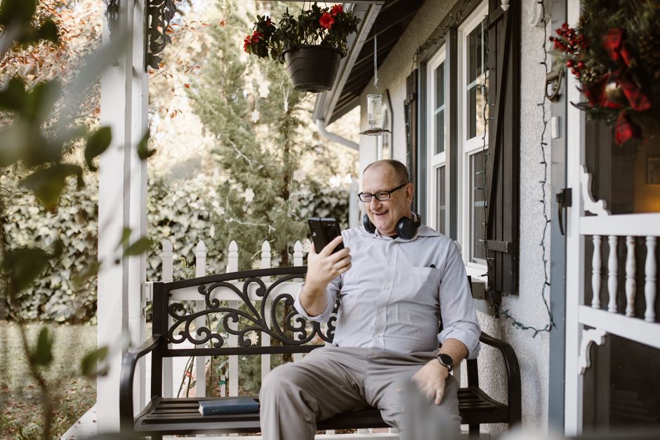 Senior man using phone and headphones for video call on home porch.
