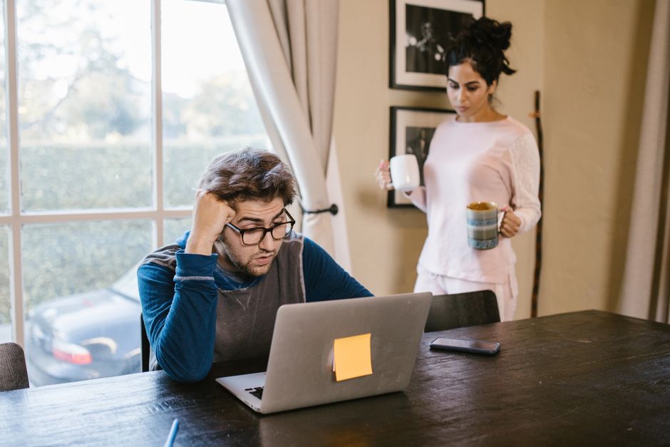 A man appears stressed working on a laptop at home while a woman brings him a drink.