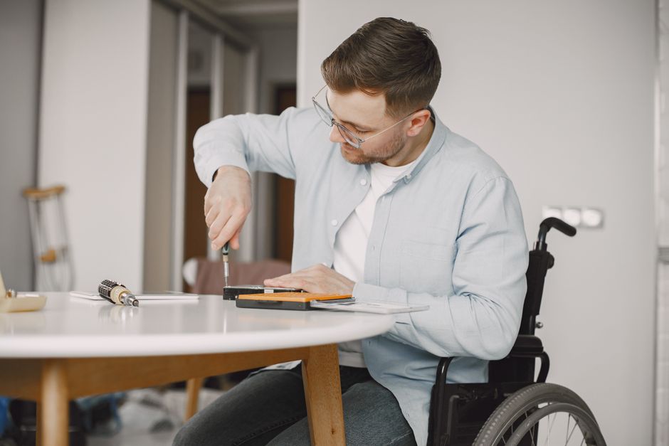 A man in a wheelchair uses a screwdriver at a home desk, engaging in repair work.