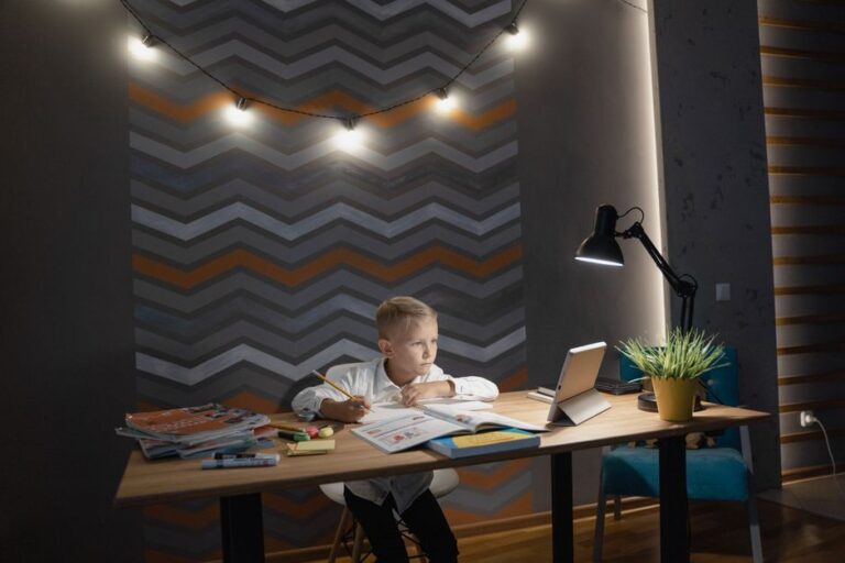 A young boy studies at a desk with a tablet, surrounded by books and well-lit by a lamp.