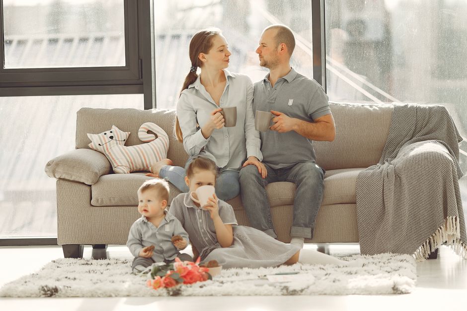 Happy family with children spending time at home while parents sitting on couch with cups of coffee and looking at each other when kids resting on floor