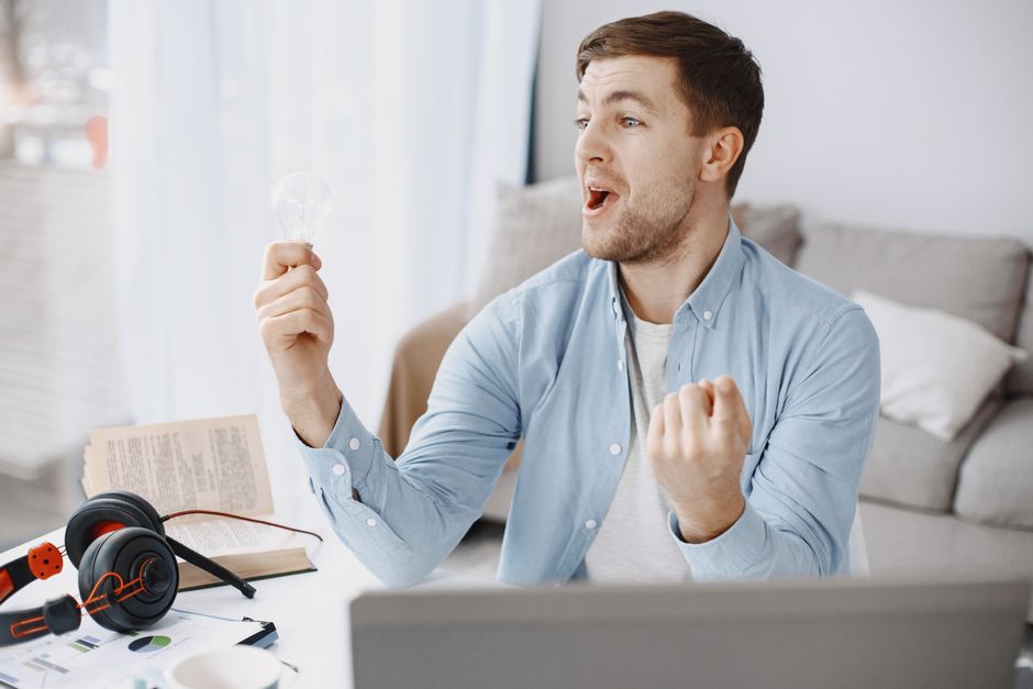 Man joyfully holding a light bulb, symbolizing creativity and innovation in a home office setting.