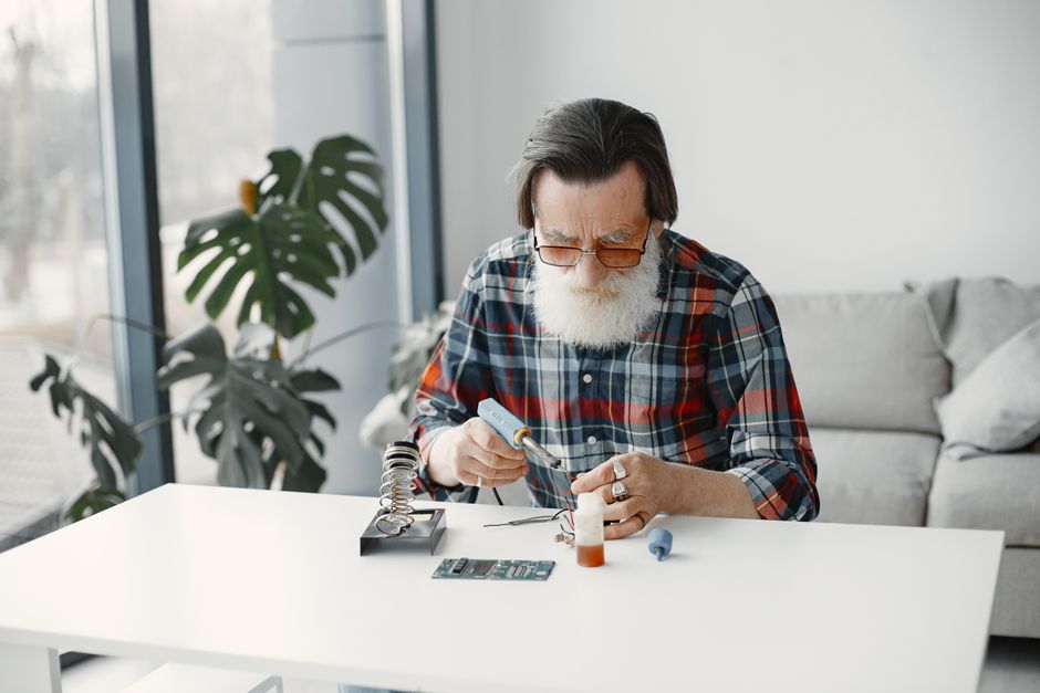Senior man with white beard in checkered shirt repairing electronics at home with tools.