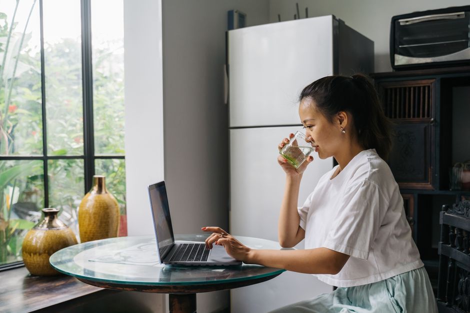 Woman sipping drink while working on a laptop at home, sitting by a window.